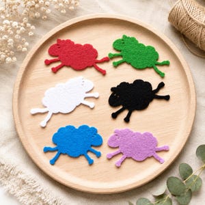 May include: Six felt sheep cutouts in red, green, white, black, blue, and purple arranged on a round wooden tray. Each sheep shape features a textured surface, a rounded body, and four thin legs, showcasing a flat, die-cut design for crafting.