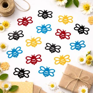 Peut inclure: Diverses découpes en forme d'abeilles colorées en rouge, noir, jaune et bleu, disposées sur une surface blanche. L'image comprend des marguerites, de la ficelle et un cadeau emballé, suggérant un thème artisanal ou de cadeaux.