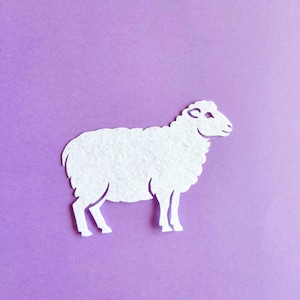 Pode incluir: Recorte branco em forma de ovelha sobre um fundo roxo liso. A ovelha é feita de um material texturizado, com um corpo fofo e um rosto sorridente. Vista de cima, a ovelha em perfil.