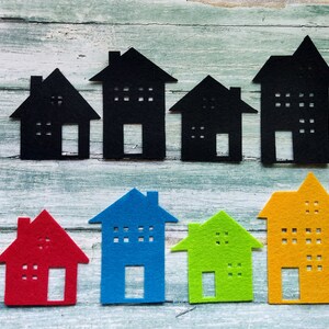 Puede incluir: Un conjunto de diez formas de casas de fieltro en varios colores, incluyendo rojo, azul, verde y amarillo. Las casas están dispuestas en una fila sobre un fondo de madera clara.