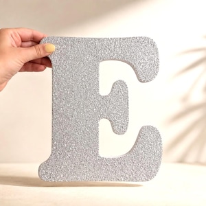 May include: A large, silver glitter-covered letter "E" is held by a hand. The letter is thick and stands out against a soft, neutral background. The letter is approximately 30 cm tall. The background has a shadow.