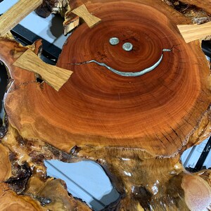 May include: A wooden slice with a natural edge, featuring a smiley face design created with silver inlay. The wood has a rich reddish-brown colour and a natural, rustic finish.