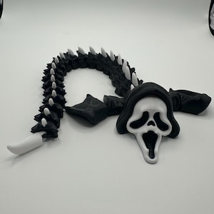 May include: A black and white articulated toy in the shape of the Ghostface mask from the Scream horror film franchise. The toy has a long, segmented body with white spikes and a white blade at the end. The mask is white with black eye and mouth details.