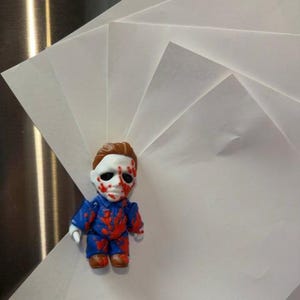 May include: A small figurine of a character with a white mask, brown hair, and a blue jumpsuit, covered in red blood-like splatters. The figurine is positioned against a fan-shaped arrangement of several blank, white rectangular sheets of paper.