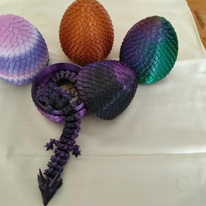 May include: Four decorative eggs with a textured, scale-like surface in purple-white, copper, and iridescent green-blue colours. One purple-black egg is open, revealing a small, articulated purple dragon figure extending outward from the interior.