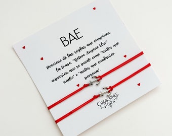 Puede incluir: Dos pulseras de hilo rojo con dijes de ancla plateados. Las pulseras están empaquetadas en una tarjeta blanca con el texto "BAE" y una definición del acrónimo en español.