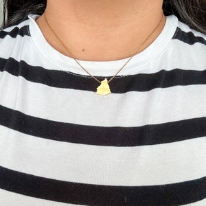 Op de afbeelding: Een gouden ketting met een klein, geometrisch hangertje. De ketting wordt gedragen door iemand die een zwart-wit gestreept shirt draagt.