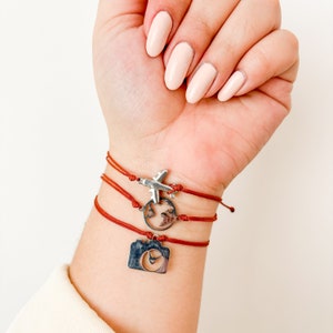 May include: A red string bracelet with three silver charms: a plane, a globe, and a camera. The bracelet is worn on a person's wrist.