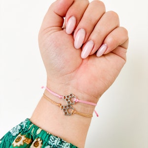 Op de afbeelding: Twee vriendschapsarmbanden met een zilveren pootprintbedel. De armbanden zijn roze en bruin en zijn om een pols gebonden.