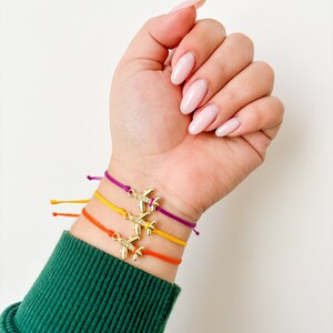 May include: Four colourful friendship bracelets with gold airplane charms. The bracelets are layered on a person's wrist.
