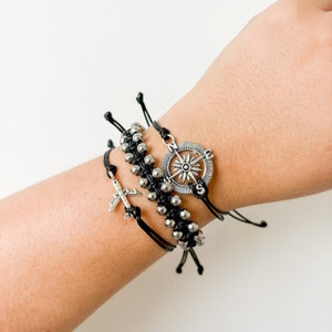 Puede incluir: Tres pulseras de cuerda negra con dijes de plata. Un dije es una brújula y el otro es un avión pequeño.
