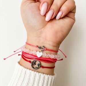 Puede incluir: Un conjunto de cuatro pulseras con hilo rosa y rojo y cuentas. Las pulseras presentan un dije de ancla de plata, un dije de corazón blanco y un dije de plata con un diseño de mapa del mundo.