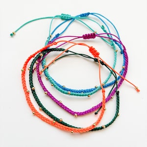 May include: A collection of colorful friendship bracelets. The bracelets are in shades of orange, green, purple, blue, and teal. Each bracelet features small gold beads and adjustable knots. The bracelets are displayed on a white surface.