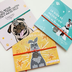 以下が含まれることがあります： 犬のイラストと足跡チャームブレスレットが描かれた3枚のカラフルなカード。カードにはパグ、シュナウザー、スペイン語のテキストが入った犬が描かれています。ブレスレットはコードに足跡チャームが付いています。