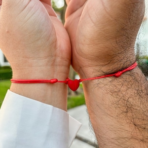 May include: Two wrists with red string bracelets tied together with a red heart charm.