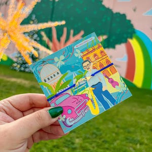 Könnte beinhalten: Ein blaues Armband mit einem goldenen Anhänger und eine Karte mit einer farbenfrohen Illustration einer Stadtszene mit einem rosa Auto, einem Mann, der Saxophon spielt, und dem Text "@creaciones_lyk".