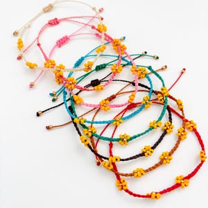 May include: A collection of adjustable cord bracelets in various colours including red, pink, teal, brown, and cream. Each bracelet features small yellow flower-shaped beads and tiny metallic gold beads spaced along the cord with sliding knots.