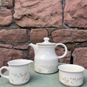 May include: A white ceramic tea set with a floral pattern. The set comprises a teapot, a creamer jug, and a sugar bowl. The floral design features pink flowers and green leaves. The set is displayed on a green surface against a brick wall.