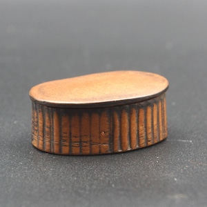 May include: A small, oval wooden box with a lid. The box is a warm brown colour, with vertical lines carved into the sides. The lid is smooth and rounded, and the box appears to be made of a single piece of wood.