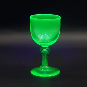 May include: A single, luminous green glass goblet with a rounded bowl and a detailed stem. The glass is set against a dark background, highlighting its vibrant colour and elegant design. The goblet is likely made of a type of glass that glows under certain light conditions.