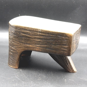May include: A small, handcrafted wooden stool with a rectangular seat and three legs. The wood has a textured, carved appearance and a dark brown finish, with a lighter-colored top. The stool is designed for decorative or functional use.
