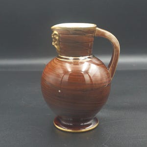 May include: A small ceramic jug with a glossy brown finish featuring a wood grain pattern. The jug has a rounded body, a narrow neck, a single handle on the right side, and a gold-coloured rim along the top edge and the base.