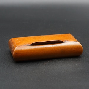 May include: A small, rectangular wooden box with a curved top and a recessed opening. The box is a warm, medium brown colour and appears to be made of a single piece of wood. The box is set against a dark background.