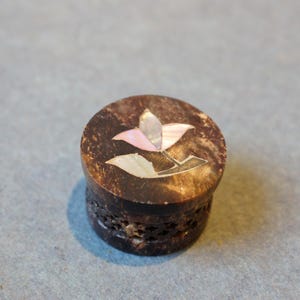 May include: A small, round, wooden box with a floral design. The box is made of dark wood and has a light pink and white flower design on the top. The box has a carved design around the edge.