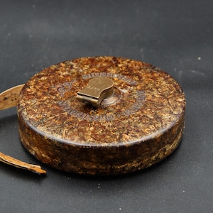 Op de afbeelding: Een vintage bruin en goud meetlint met een messing opwindmechanisme. De tape komt uit een cirkelvormige behuizing met een textuur. De behuizing heeft reliëftekst rond de messing opwindsleutel.