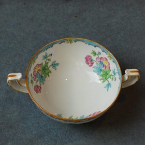May include: A white porcelain soup bowl with a floral design in shades of pink, blue, and green. The bowl has two handles and a gold rim.