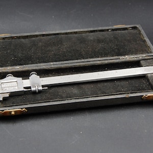Könnte beinhalten: Ein Vintage-Metall-Messschieber-Werkzeug, offen in seinem schwarzen Etui. Der Messschieber ist silberfarben mit einer Messskala. Das Etui ist rechteckig mit messingfarbenen Verschlüssen und einem schwarzen Samtinnenraum. Das Werkzeug dient zum präzisen Messen.