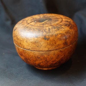 Puede incluir: Una caja redonda de madera con tapa. La caja es de color marrón claro cálido con marcas marrón oscuro y negro. La tapa tiene un diseño circular tallado en la parte superior. La caja está sobre un fondo oscuro.