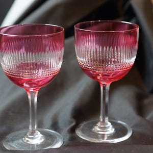 May include: Two clear glass wine glasses with a pink tint and a cut glass design. The glasses are on a black surface.