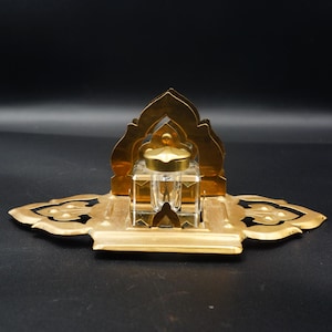 Pode incluir: Um conjunto de tinteiro vintage com um frasco de tinta de vidro transparente e uma base de metal dourado. A base tem um design ornamentado com um arco decorativo e uma bandeja. O tinteiro é provavelmente do início do século XX.