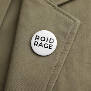 Pode incluir: Um botão branco com as palavras "ROID RAGE" em letras maiúsculas pretas em negrito. O botão está preso a um casaco cáqui. O botão é redondo e tem um acabamento brilhante.