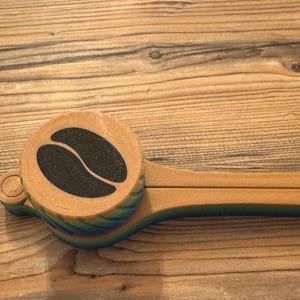 Könnte beinhalten: Ein hölzerner Kaffeelöffel mit rundem Griff und einem schwarzen Kaffeebohnen-Design. Der Löffel hat einen langen, flachen Griff und ist mit grünen und blauen Akzenten bemalt. Der Artikel befindet sich auf einer Holzoberfläche.