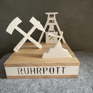 Könnte beinhalten: Ein dekoratives Holzstück mit weißen ausgeschnittenen Formen von gekreuzten Hämmern mit dem Text "GLÜCK AUF", einem Bergwerksschacht und einer Bergkette. Die Basis ist aus Holz mit dem Wort "RUHRPOTT" ausgeschnitten.