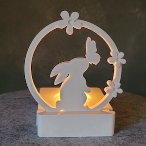 Könnte beinhalten: Weißer Kerzenhalter aus Keramik mit einem Ausschnitt in Form eines Hasen und eines Schmetterlings. Der Halter hat eine kleine, runde Öffnung für ein Teelicht.