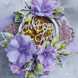 May include: A handcrafted lavender-colored box decorated with purple fabric flowers, green leaves, and a gold glitter-filled center. The center features the text "Mażę Iono" in white script. Decorative paper cutouts and pearls add detail.