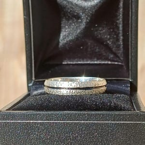 Könnte beinhalten: Ein silberner Ring mit gehämmertem Muster und einem schwarzen Band, präsentiert in einer schwarzen Schmuckschatulle. Die Oberfläche des Rings reflektiert das Licht und betont seinen metallischen Glanz. Der Ring ist ein Schmuckstück.