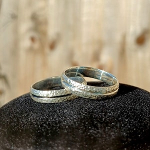 Könnte beinhalten: Zwei silberne Ringe mit gehämmerten Texturen werden auf einer schwarzen Oberfläche gezeigt. Die Ringe haben ein schlichtes, elegantes Design mit einem strukturierten Band und einem glatten, dünnen Band. Der Hintergrund ist eine verschwommene Holzoberfläche.