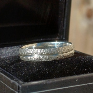 Könnte beinhalten: Ein silberner Ring mit gehämmertem Muster, präsentiert in einer schwarzen Ringschatulle. Der Ring hat ein strukturiertes Außenband und ein glattes, poliertes Innenband. Das Design ist schlicht und elegant.