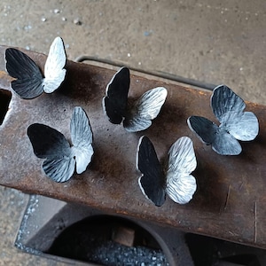 Op de afbeelding: Zes handgemaakte metalen vlindersculpturen met getextureerde vleugels liggen op een donker, verweerd oppervlak. De vlinders zijn in verschillende tinten grijs en zwart, wat gedetailleerd vakmanschap laat zien. De vleugels zijn open, wat de indruk van vlucht geeft.