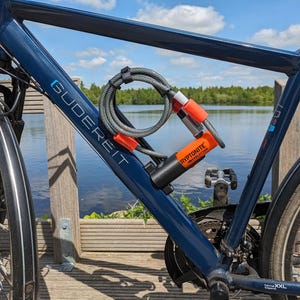 Könnte beinhalten: Ein blaues Fahrrad mit einem Kryptonite-Schloss am Rahmen befestigt. Das Schloss, mit einem grauen Kabel, ist schwarz und orange. Auf dem Fahrradrahmen steht das Wort "Gudereit". Im Hintergrund ein See und Bäume.