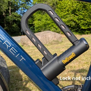 Könnte beinhalten: Schwarzes U-förmiges Fahrradschloss an einem blauen Fahrradrahmen befestigt. Das Schloss trägt das Wort "ONGUARD" in Gelb. Der Text "Lock not included" befindet sich am unteren Bildrand. Das Fahrrad befindet sich im Freien mit Bäumen im Hintergrund.