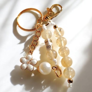 Puede incluir: Llavero dorado con un dije de flor, una cadena y un cierre de mosquetón. El llavero presenta una hebra de cuentas de color amarillo y una perla grande. La flor tiene pétalos blancos y un centro dorado.