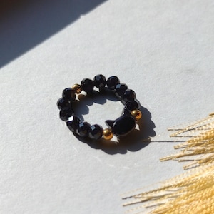 May include: A black beaded ring with a cat-shaped charm. The ring features faceted black beads and small gold-colored beads. The cat charm is also black. The ring is displayed on a white surface with a shadow.