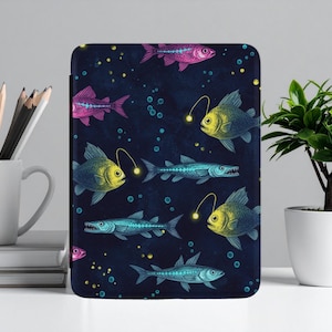 Könnte beinhalten: Eine dunkelblaue Tablet-Hülle mit einem sich wiederholenden Muster aus bunten Fischen. Das Design umfasst gelbe, blaue und rosa Fische mit leuchtenden Akzenten, vor einem Hintergrund aus Blasen und Sternen.