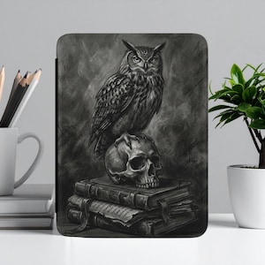May include: A black and white illustration of an owl perched on a skull, atop a stack of books. The artwork is displayed on a tablet case. A white mug with pencils and a small potted plant are also visible.