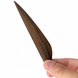 Op de afbeelding: Een donkerbruin houten vouwbeen met een puntige uiteinde en een gebogen rand. Het oppervlak van het hout toont een natuurlijk nerfpatroon en het woord "BONEFOLDER" is in kleine, duidelijke hoofdletters aan de zijkant van het gereedschap gedrukt.